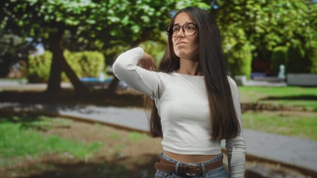Woman with glasses hand behind head touching hair on street path in park; thoughtful doubt reflection.