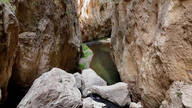 High angle view of a narrow gorge with a winding stream flowing between steep, eroded canyon walls and large boulders on a sunny day
