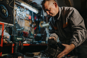 A skilled technician in a gray uniform works at a cluttered workshop, examining and assembling a component with a cordless drill. The scene conveys focus, hands-on craft, and industry atmosphere.