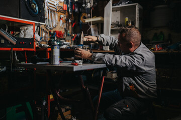 A focused mechanic works at a cluttered workshop bench, using a drill and hammer among tools and parts. Dim lighting and organized chaos capture hands-on repair work and craftsmanship.