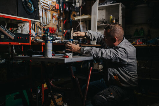 A focused mechanic works at a cluttered workshop bench, using a drill and hammer among tools and parts. Dim lighting and organized chaos capture hands-on repair work and craftsmanship.