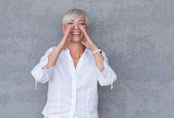 Amused woman yelling, against gray background - wall. Happy middle age woman holding hands near his mouth