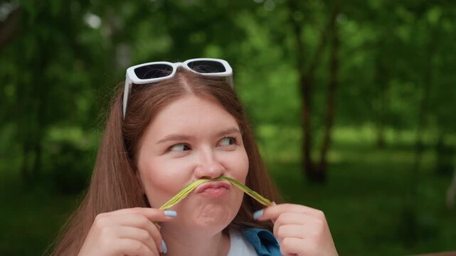 Close up of young woman sitting outdoors holding colorful thread across face pretending mustache while making funny playful expression, laughing cheerfully in green park