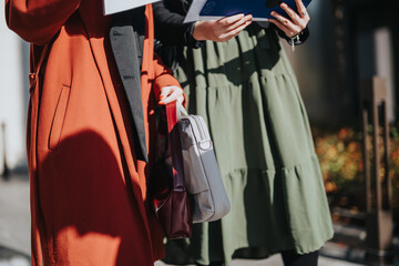 Fototapeta premium Two professionals stand outdoors, holding tablets and folders, wearing bold coats and carrying bags. The scene conveys collaboration, urban style, and active business engagement in the city.
