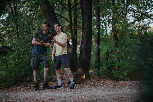 Two men in casual outdoor gear stand by a tree in a wooded park, looking at a smartphone map. Backpacks rest nearby as they plan their hike together.