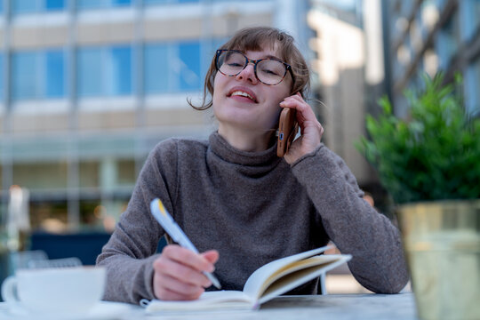 Young woman enjoying a sunny day while talking on the phone and taking notes at an outdoor cafe