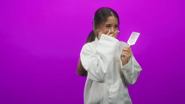Young woman chef wearing white coat holding a small grater, hand to mouth gesture while smiling against purple studio backdrop; playful amusement.