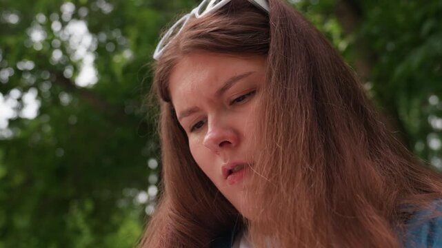 Close up focused lady with glasses on head working outdoors, concentrated face framed by brown hair, intent gaze while managing craft task in park under soft daylight