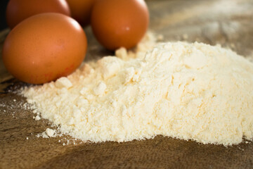 Close-up top view of brown eggs and a mound of flour on a dark wooden board. Dramatic lighting highlights textures, creating a warm, rustic, emotional atmosphere perfect for baking themes.