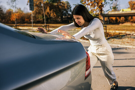 A young woman trying to push her car that won&rsquo;t start, struggling on the roadside, daylight, casual everyday moment.