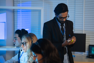 Business manager takes notes while overseeing a team of customer service agents working late in command center or call center office. high-pressure operation involving professional communication