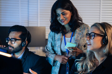 Smiling female manager provides coaching to a customer service agent wearing a headset in a dark office. Corporate training, IT helpdesk, professional assistance, and late night call center