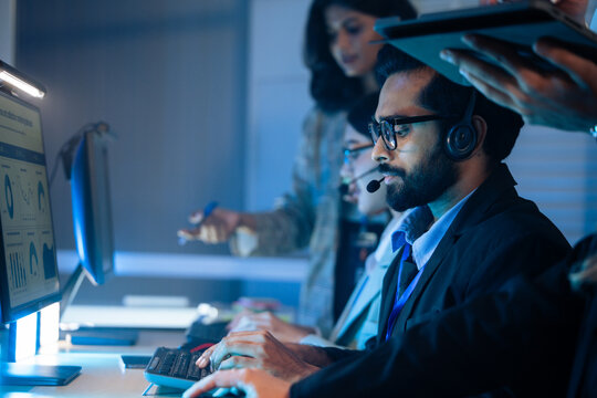 Customer service agent wears a headset talking to a client in a dark call center. Support, IT helpdesk staff, and late night corporate operations and assistance telemarketing - Powered by Adobe