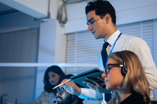 Serious manager uses a tablet to mentor customer service agents wearing headsets in a control room. Corporate training, IT helpdesk supervision, professional support staff, and late night call center 