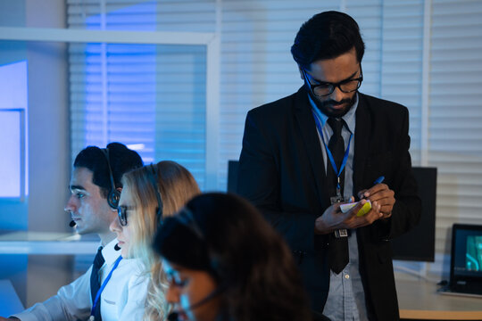 Business manager takes notes while overseeing a team of customer service agents working late in command center or call center office. high-pressure operation involving professional communication - Powered by Adobe