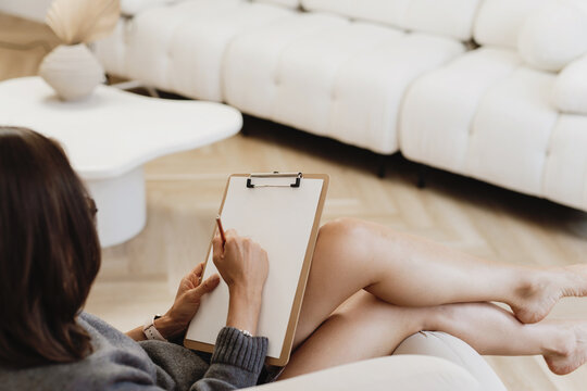 A woman relaxing on a modern sofa while writing on a blank clipboard.