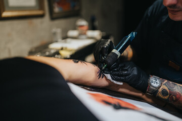 A tattoo artist wearing black gloves uses a tattoo machine to ink a design on a client's forearm, capturing a precise, intimate moment of body art in a studio setting.