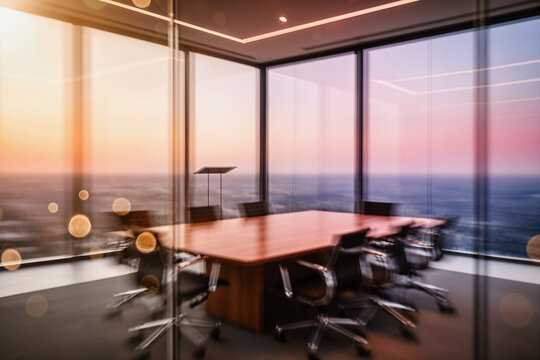 A modern conference room with a wooden table and black chairs, featuring large windows overlooking a sunset over the ocean, captured with a shallow depth of field and bokeh effects.