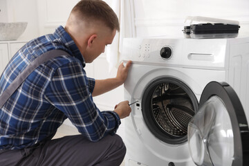 Repairman in uniform fixing broken washing machine with screwdriver at home