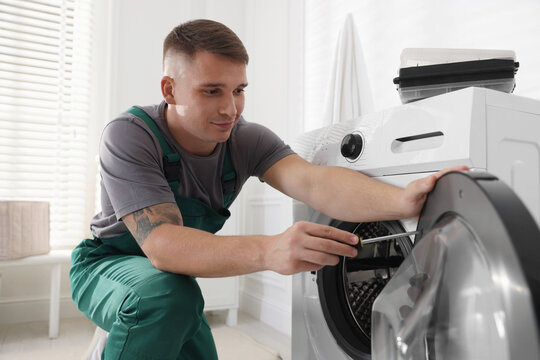 Repairman in uniform fixing broken washing machine with screwdriver at home