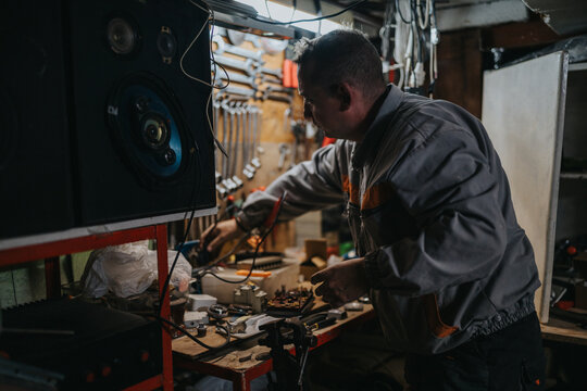 A skilled technician repairs electronic devices in a busy workshop filled with tools, wires, and spare parts. The focused worker uses hand tools to assemble components on a workbench.