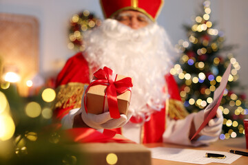 Saint Nicholas with gift and letter at desk in room decorated for Christmas, selective focus