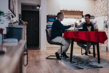 Two men sit at a kitchen table with a red tablecloth, raising glasses in a cheerful toast. They enjoy a relaxed evening at home, sharing food, conversation, and friends company.