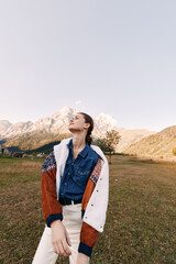 Fototapeta premium Woman standing in field with mountains in background, wearing patterned sweater and denim shirt, portrait style fashion shot in autumn light, casual model outdoors.