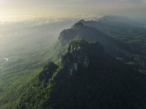 Aerial view of the majestic, cloud-kissed mountain peaks rise above a verdant forest, a symphony of green and grey under a soft, diffused light, Tambon Wichet Nakhon, Chang Wat Lampang, Thailand.