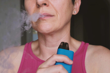 Close-up of middle-aged woman exhaling vapor while holding a blue vape device, illustrating modern nicotine consumption habit