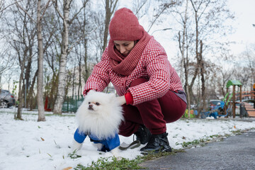 Woman crouches to correct and secure the harness on her white Spitz, showing basic training and handling during a winter walk in a residential city area.