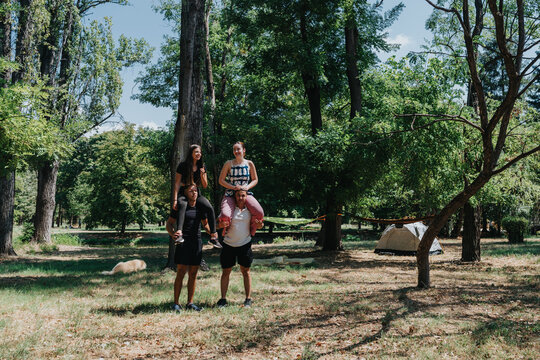 A cheerful family spends time outdoors at a wooded campground. Parents carry their children on their shoulders while others stand nearby, with a tent set among trees under a bright sky. - Powered by Adobe