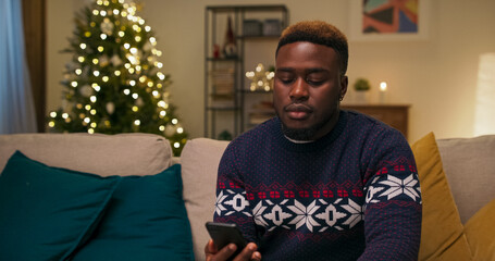 A young man of multi-ethnic background sits on a sofa in the living room. His mother called to wish...