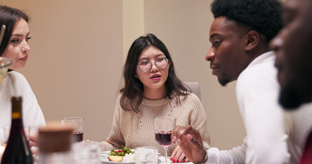 Sitting at a table, a girl of Asian descent in glasses explains the method of preparation of a dish to a multi-ethnic boy with a beard. The brunette tells the guy about the food.