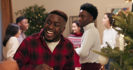 A multi-ethnic guy holds a box of decorations as he and his girlfriend a couple in love stand near a Christmas tree and hang decorations at a Christmas party. They are talking and smiling while the