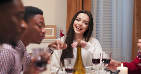 Christmas Eve. Friends are sitting at the festive table, talking and eating. Attractive young girl with dark long hair puts cutlery, looks at camera, rests head on hand and smiles.