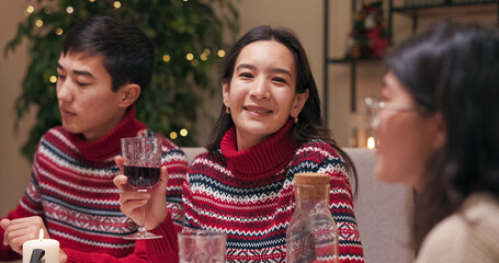 An attractive girl with dark long hair is sitting with friends at the festive table. She is wearing a red warm sweater and has gold earrings. The woman holds a glass of wine, looks at the camera, and