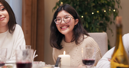 Sitting at the table and celebrating Christmas, young people are smiling and having fun. A girl of Asian descent in glasses and with black hair is talking with friends, while also looking at the