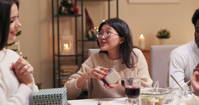 A multi-ethnic girl in glasses opens a gift and is satisfied while sitting at the festive table with friends. The brunette with long hair gave the gift, and both girls are happy and smiling.