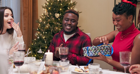 A multi-ethnic guy in a red festive shirt gives a gift to his girlfriend, a girl in a red turtleneck, who is happy and unwraps it. Friends rejoice for the couple in love and smile as they sit at the