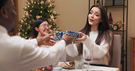 A guy gives a gift to his girlfriend during a party. The brunette in a white sweater unties the ribbon, opens the box, and is pleasantly surprised. She shows it to friends, thanks her boyfriend, and
