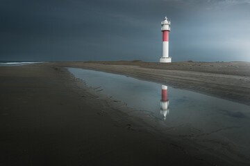 Red and white Fangar lighthouse reflecting on a wet beach under a dark stormy sky at the Ebro Delta