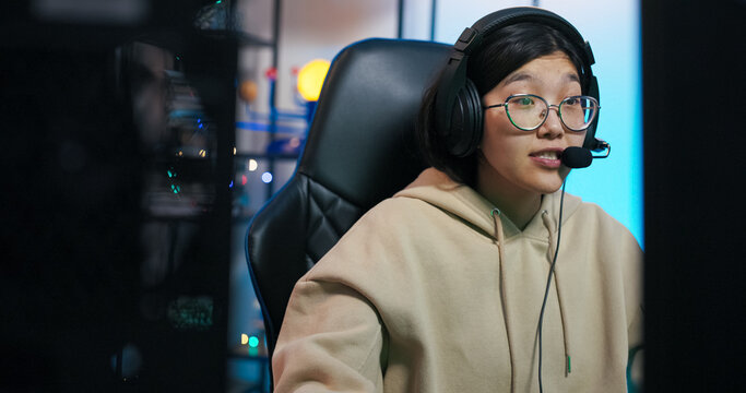 Young woman sits in room lit by blue led lights in front of computer gamer puts on headset improves microphone, greets friends met on internet in an online game, a girl prepares to play