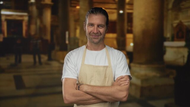 Blond man with arms crossed wearing linen beige apron inside ornate catholic church building; serenity.