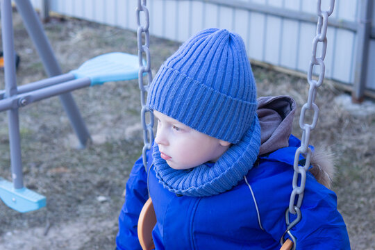 Little boy dressed in a warm blue hat and jacket, sitting on a swing in a park or backyard looking sad and thoughtful. Child emotions and childhood concept. - Powered by Adobe