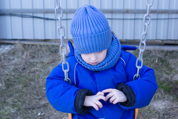 Close up of a small kid in a blue hat and warm suit, making a heart shape with hands while sitting on a swing. Concept of love and care.