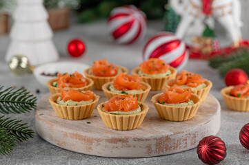 Festive Christmas canapes. Mini tartlets with creamy avocado filling and smoked salmon, garnished with herbs on a rustic wooden board on a gray concrete background. Selective focus. Copy space.