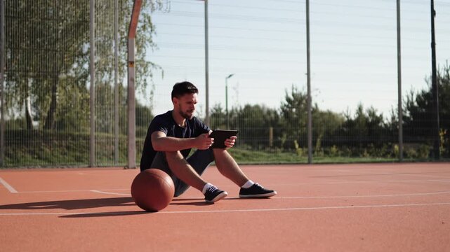 Caucasian man sits on basketball court near ball, looking at tablet and gesturing frustratedly. Portrait of emotional fan reacting to loss and setback on sunny day. Watch match online from anywhere.