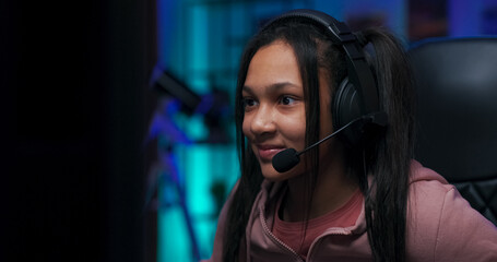 Focused teenage girl in headset plays video games in LED-lit room. She concentrates while passing another level on the computer, immersed in her virtual world with purple lights glowing.