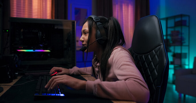 A professional gaming girl sits in front of her screen, speaking to team members through a microphone. Purple LED lights fill the room while she plays with focus and joy. - Powered by Adobe